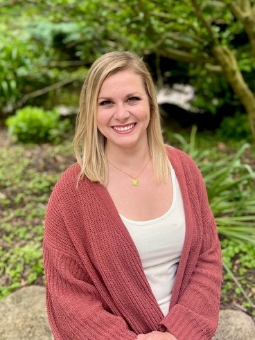 White woman posing with greenery in background.