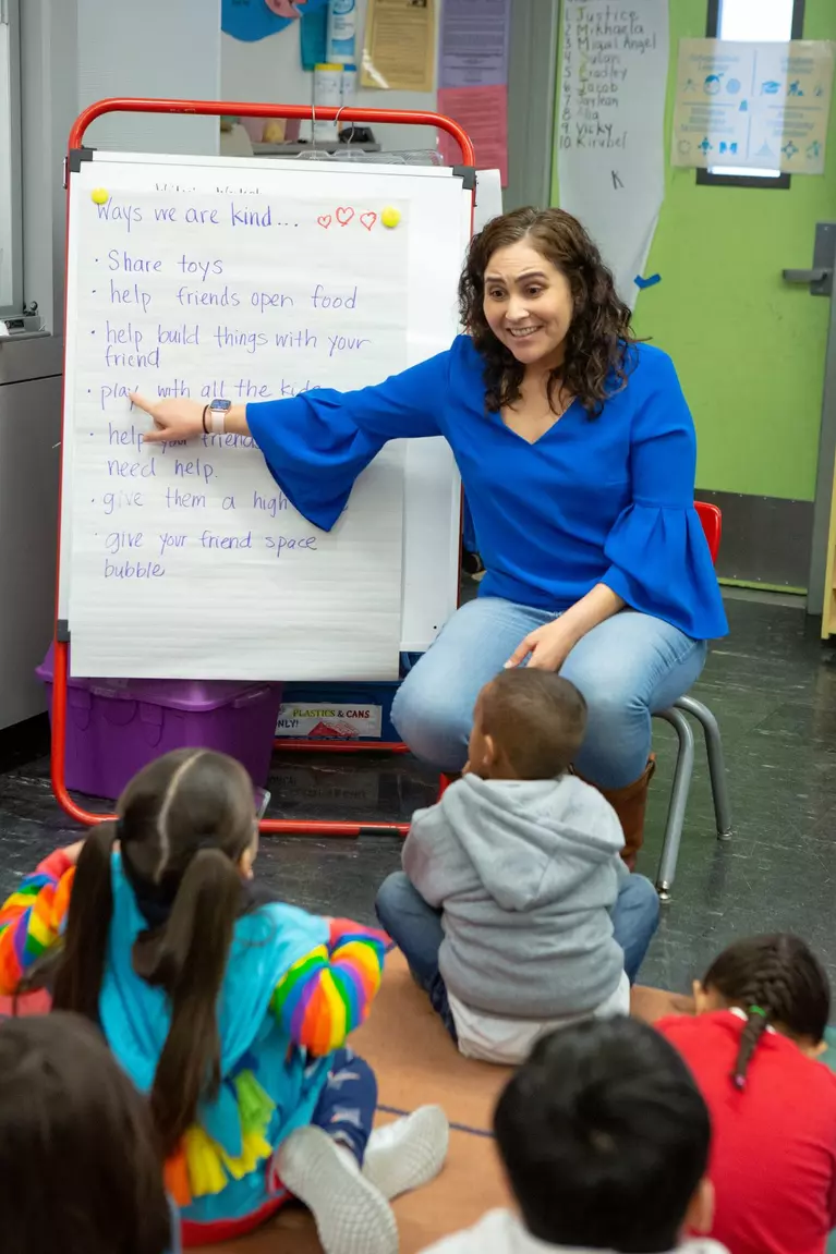 Teacher pointing to white paper flip board with preschool children looking up at her while sitting on the floor.