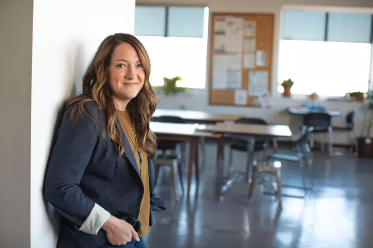 Young student teacher leaning against a wall with empty classroom in the background.